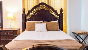 A neatly made bed with a dark wooden headboard, a bedside table with a lamp, and a small folding chair.