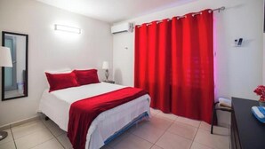Quarto de hotel com cama, escrivaninha, cadeira, espelho, abajur e cortina vermelha.