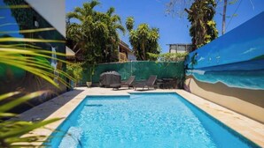 Uma piscina com espreguiçadeiras e uma área de churrasco coberta, cercada por vegetação e um céu claro.