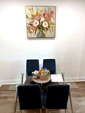 Uma área de refeições com mesa de vidro, duas cadeiras azuis, um vaso com flores e um menu sobre a mesa.