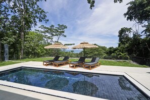 Piscina con sillas de descanso y sombrillas de paja, rodeada de vegetación y un cielo despejado.