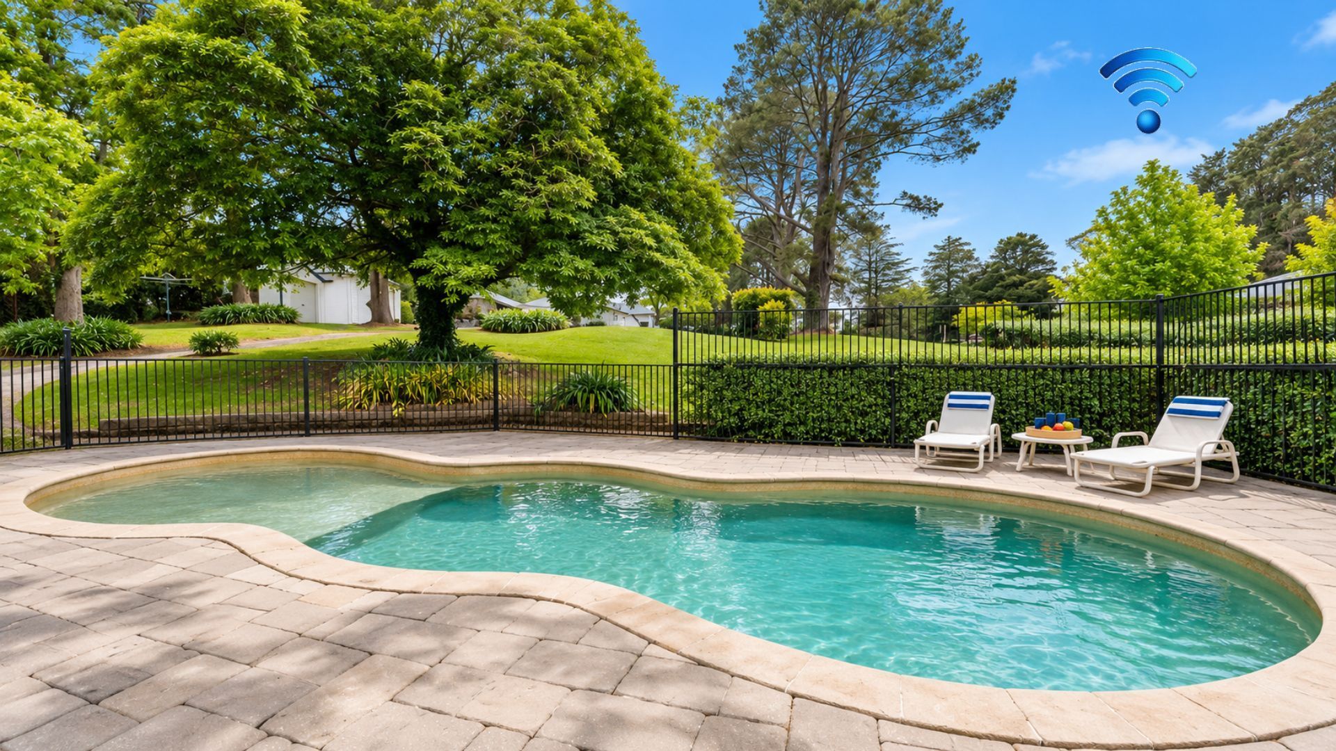 A swimming pool with a curved edge, surrounded by a paved deck. There are two lounge chairs and a small table with drinks.