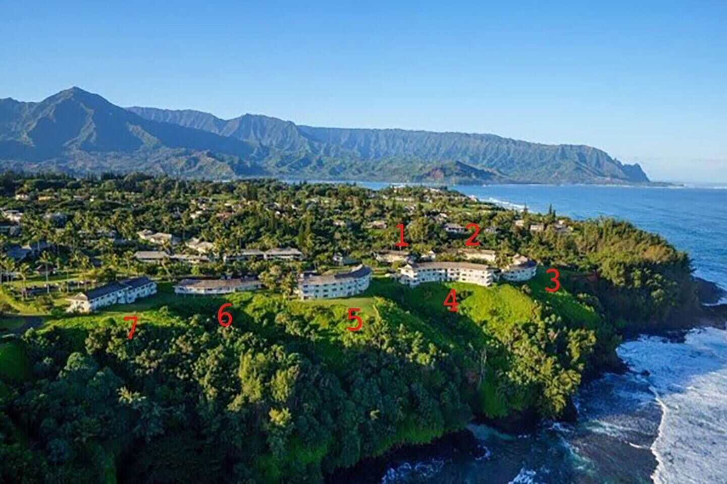 An aerial view of a coastal resort with numbered buildings, lush greenery, and a mountainous backdrop.