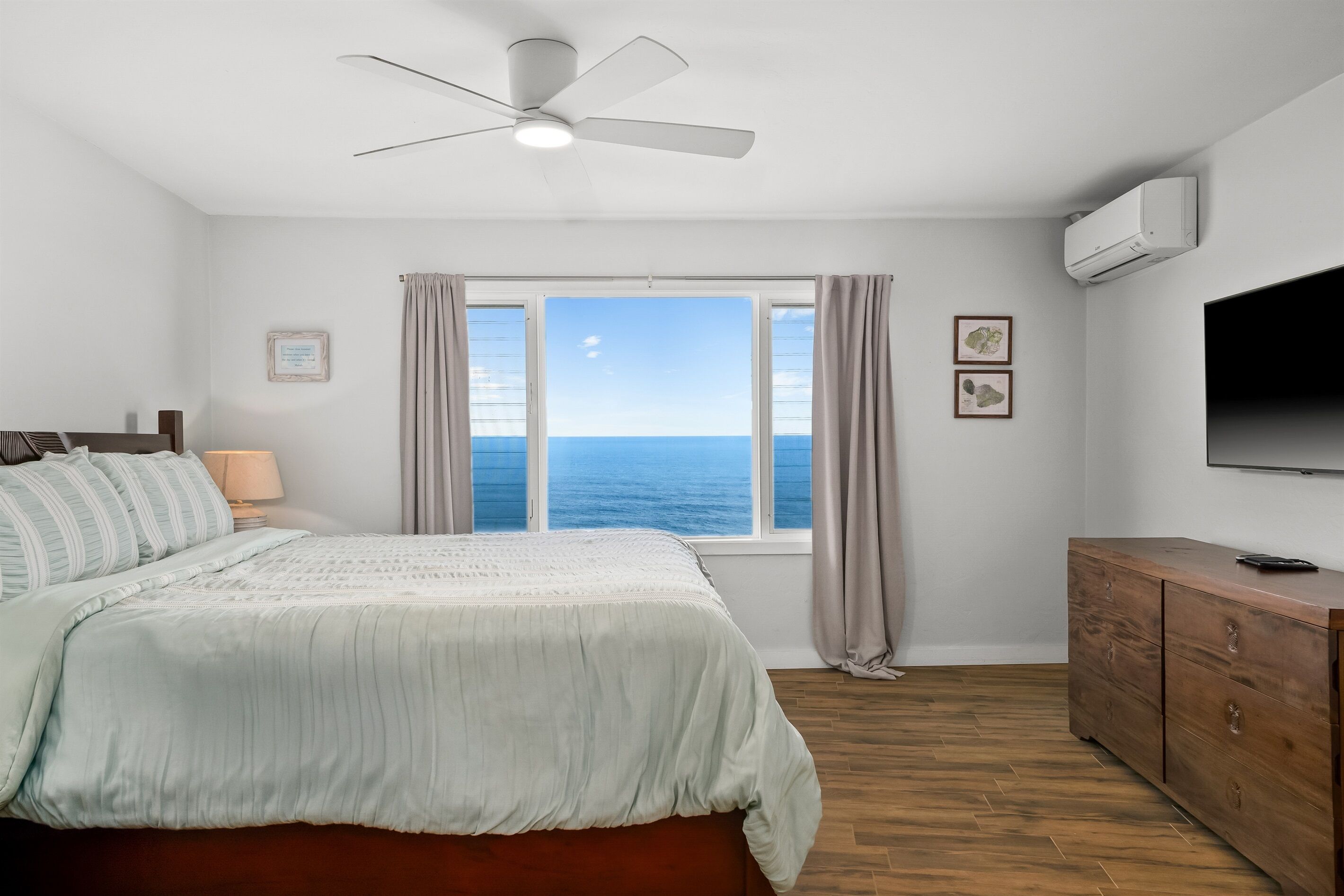 Un dormitorio con cama, ventilador de techo, ventana con vista al mar, cómoda de madera y televisor de pantalla plana.