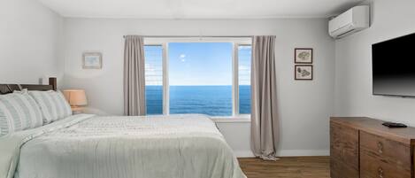 Un dormitorio con cama, ventilador de techo, ventana con vista al mar, cómoda de madera y televisor de pantalla plana.