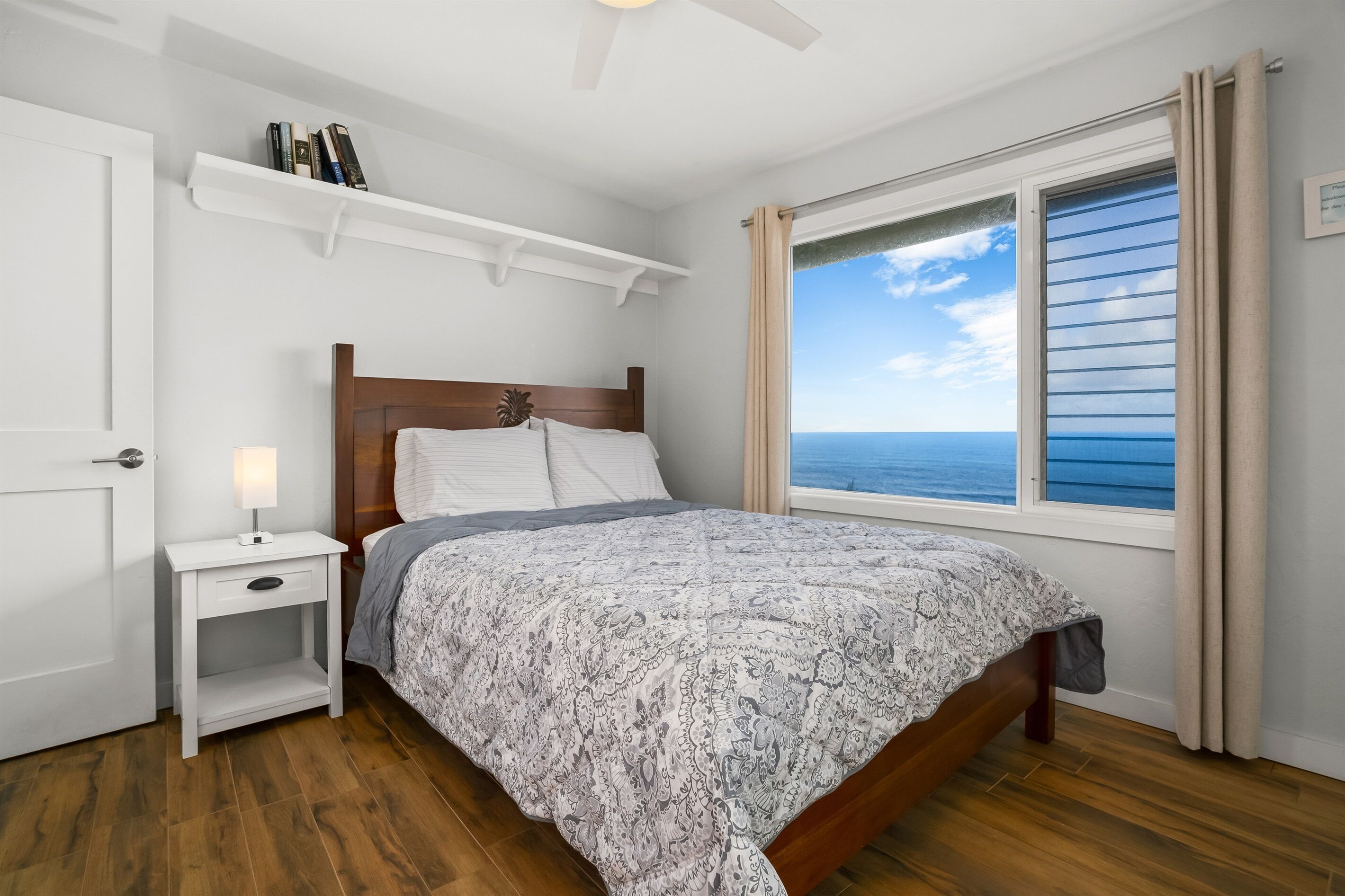 Un dormitorio con una cama de madera, una mesita de noche con una lámpara, un ventanal con vistas al océano y una estantería con libros.