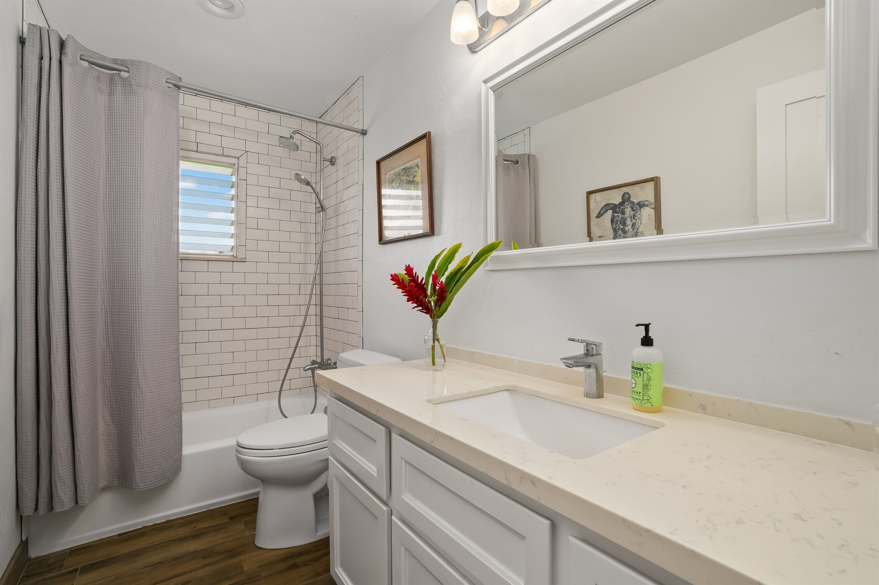 A modern bathroom with a white bathtub, toilet, and sink. There is a shower curtain, a framed picture on the wall, and a vase with flowers on the sink counter.