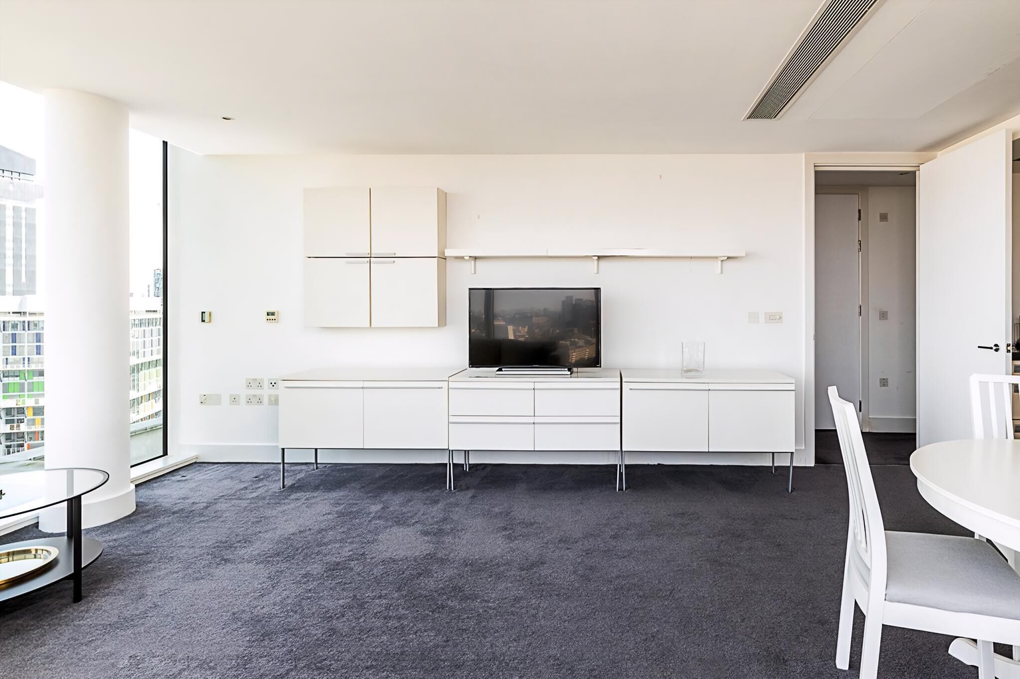 A modern living room with a large flat-screen TV mounted on a white cabinet, a glass coffee table, and a white dining table with chairs.