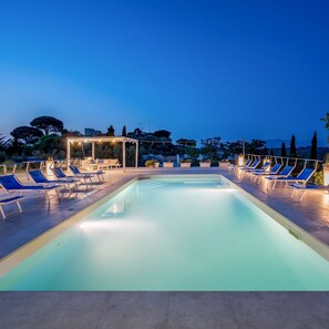 Una piscina rectangular con sillas reclinables azules a su alrededor, una pérgola con luces colgantes y vistas a árboles y edificios al fondo.