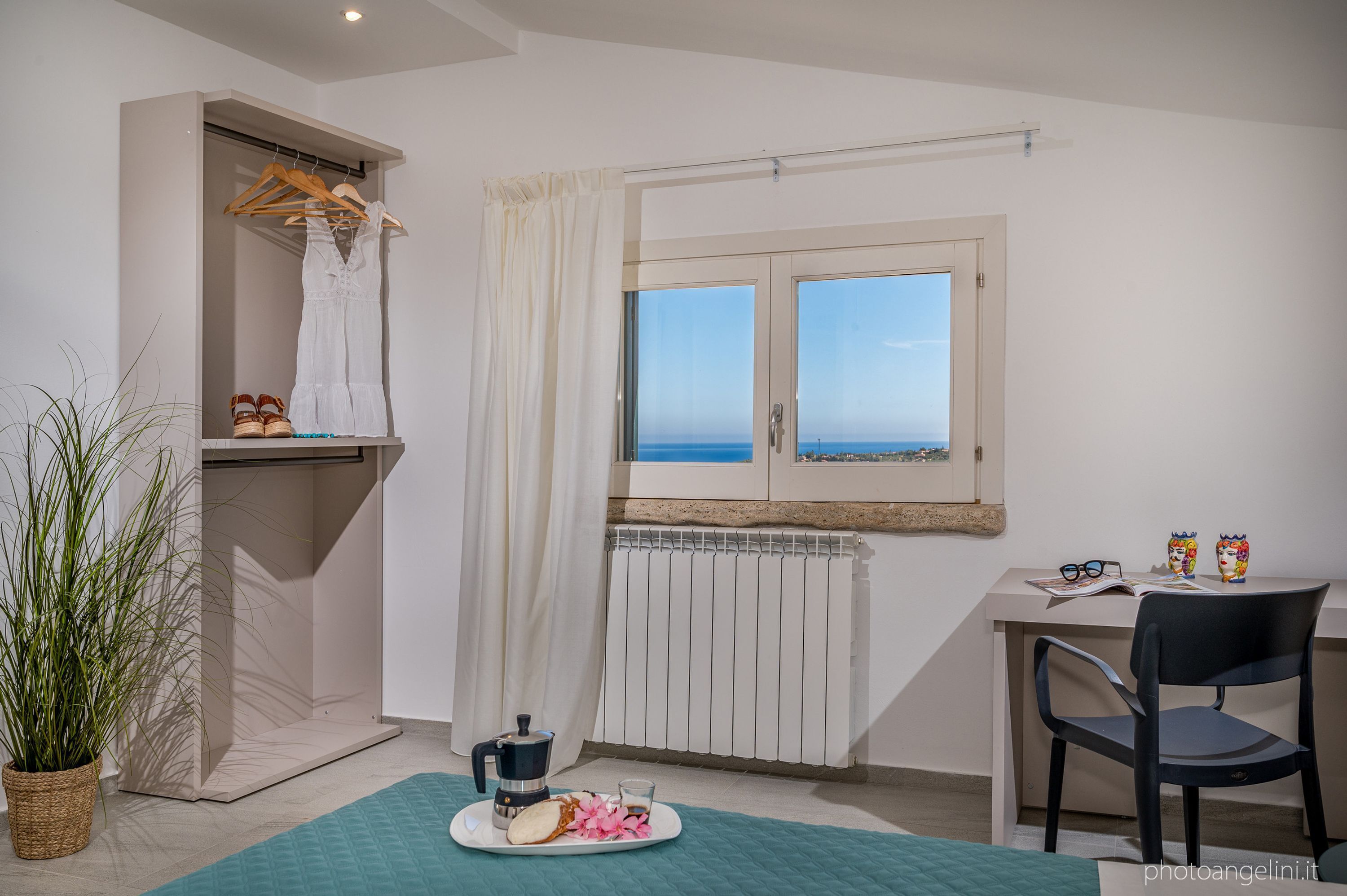 Ein Zimmer mit Bett, Schreibtisch, Stuhl, Heizkörper, Fenster mit Meerblick, einer Schrankwand und einer Zimmerpflanze.