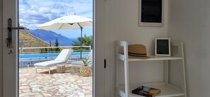 A room with a stone floor, a white chair, a small table, a potted plant, a white shelf with a hat and a framed picture, and a view of a pool and mountains through a window. - Villa Ballerina Bianca (Castellammare del Golfo)