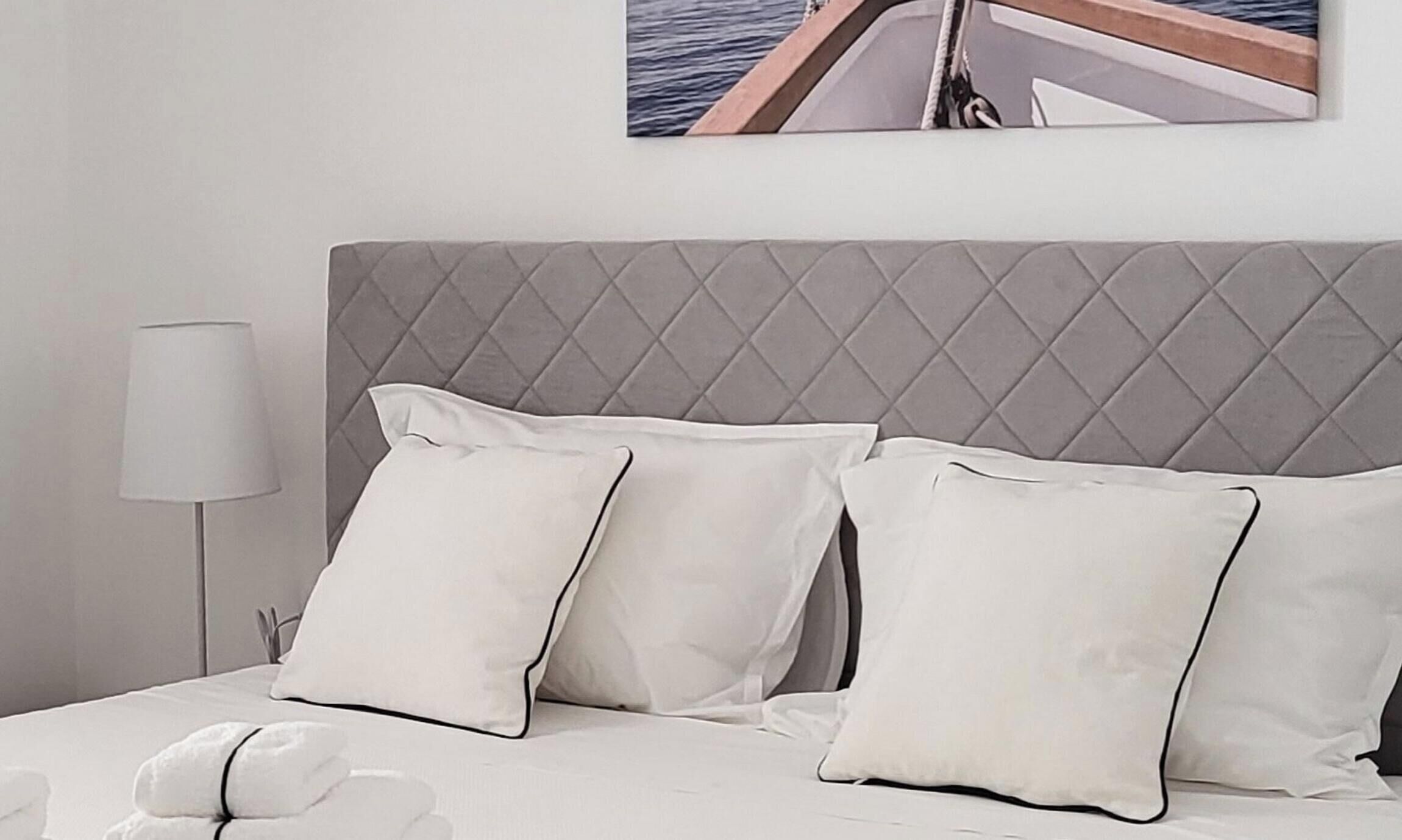 A neatly made bed with white linens and pillows, a grey headboard, and a bedside lamp.