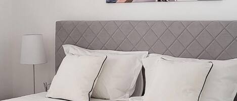 Ein ordentlich bezogenes Bett mit weißen Bettwäsche und Kissen, ein grauer Kopfteil und eine Nachttischlampe.