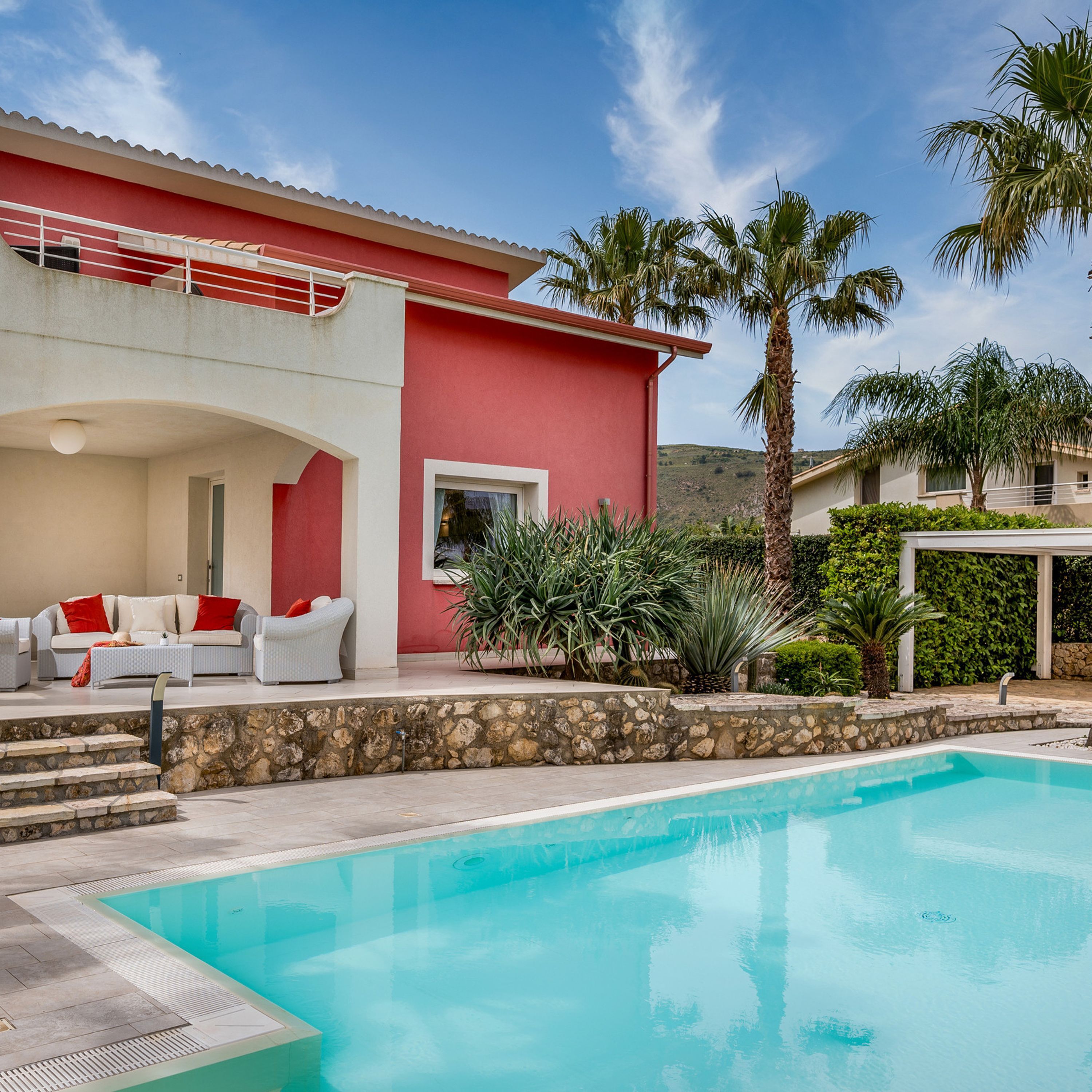 Un espace aménagé au bord de la piscine, avec un bâtiment rouge, du mobilier d’extérieur et des palmiers.