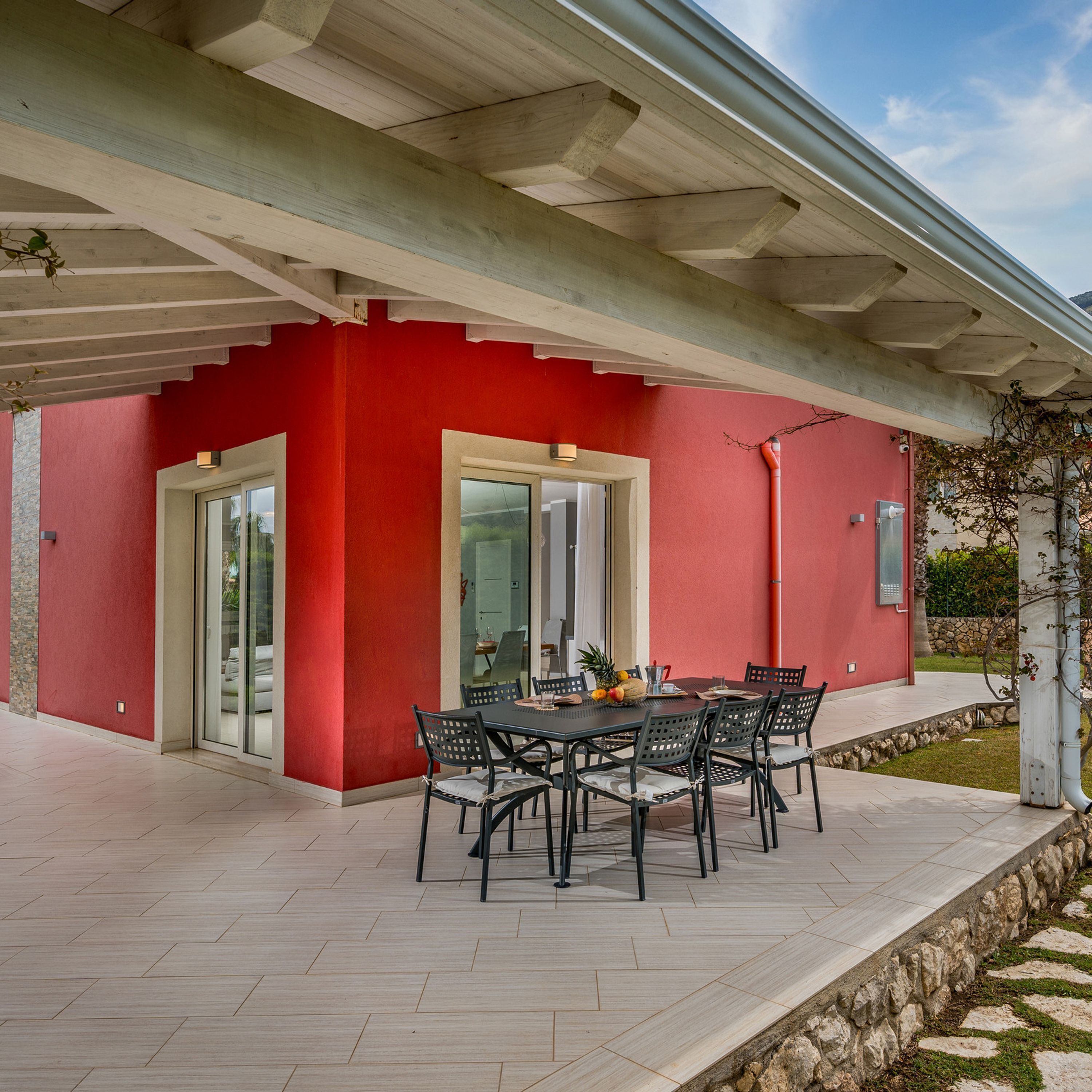 Un patio couvert avec un mur extérieur rouge, une porte d’entrée en verre et une table entourée de chaises.