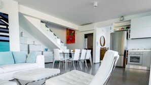 A modern living room with a white sofa, a coffee table, and a unique white chair with a curved backrest. A staircase leads to another level, and a kitchen with a refrigerator and oven is visible in the background. - Villa Majestatis in Scopello (Castellammare del Golfo)