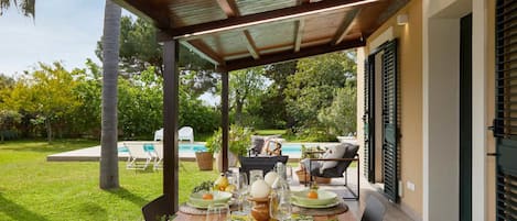 Un área de comedor al aire libre con un patio cubierto, una piscina y vistas a un jardín.
