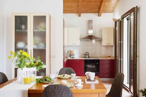 A dining area with a wooden table set for breakfast, a kitchen with red cabinets, and a sliding glass door leading to an outdoor area. - Giardino di Limoni Eco Friendly Villa (Noto)