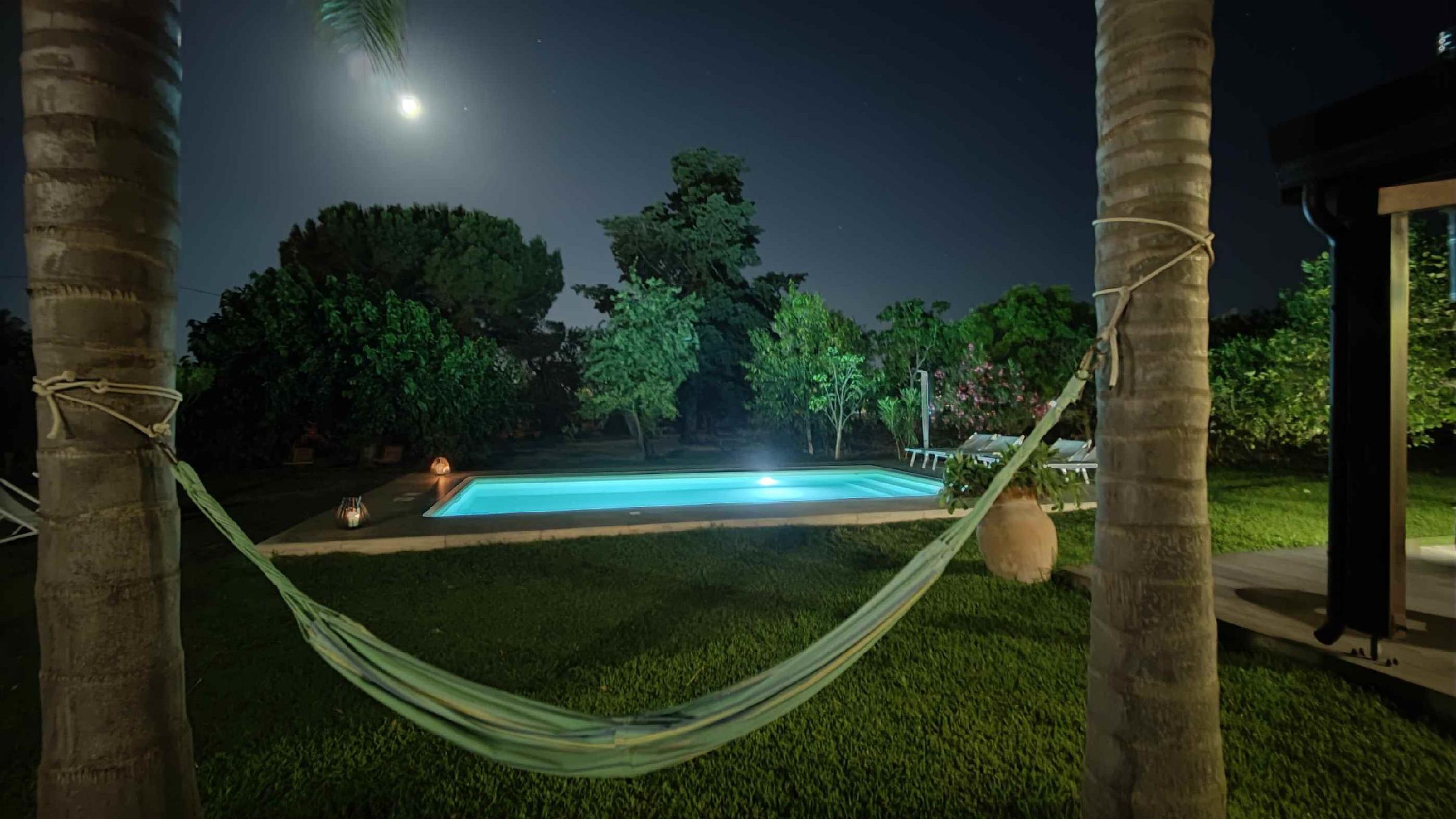 Spazio esterno illuminato di notte con piscina, amaca e alberi.
