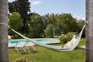Una hamaca junto a una piscina, con césped y árboles al fondo.