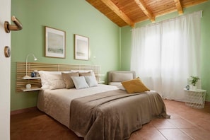 A bedroom with a bed, bedside tables, a nightstand, a lamp, a window with curtains, and a wall-mounted picture. - Giardino di Limoni Eco Friendly Villa (Noto)