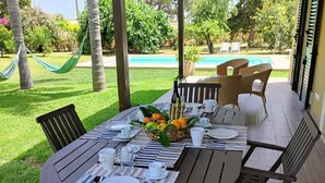 Un área de comedor al aire libre con una mesa lista para cuatro personas, rodeada de sillas, una hamaca y una piscina al fondo.