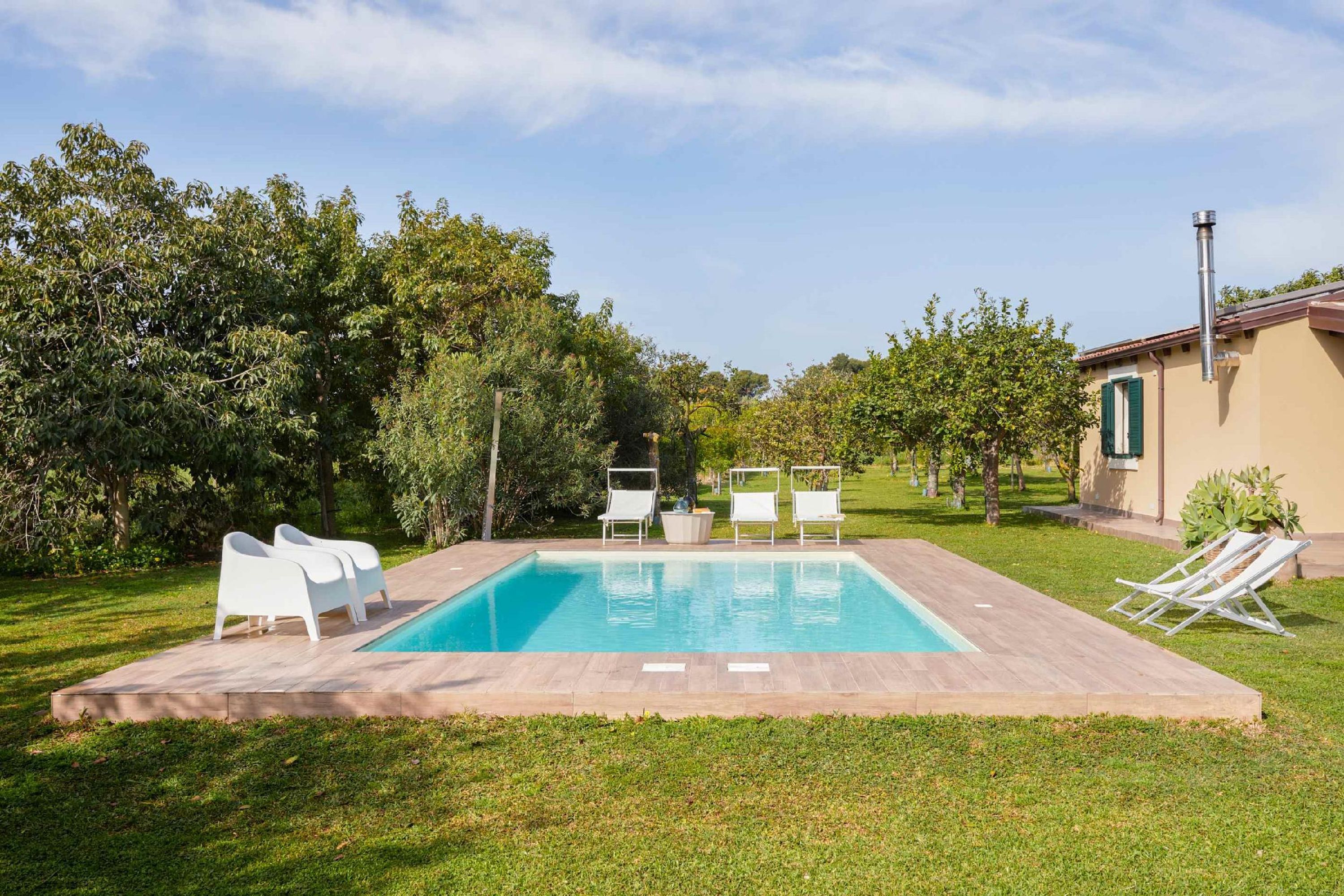 Una piscina rettangolare con bordo piastrellato, circondata da poltrone da relax bianche e un prato curato.