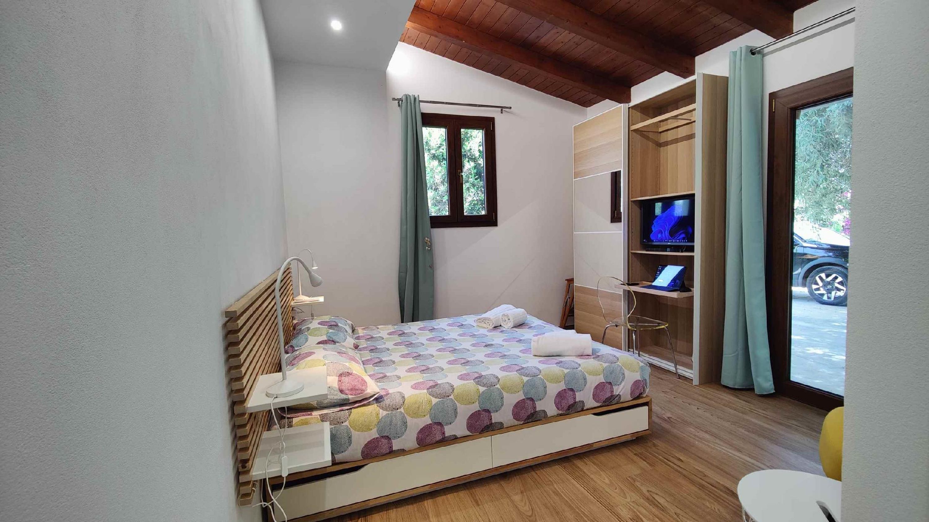A bedroom with a bed, a desk with a computer, a TV, and a window with curtains.