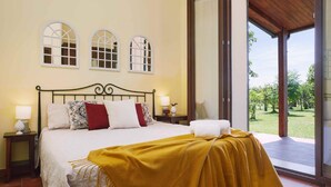 A bedroom with a bed, a wooden headboard, a window with a view of a garden, and a door leading to an outdoor area.