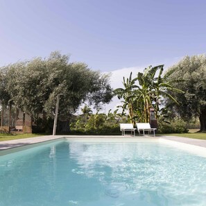 A rectangular swimming pool with clear blue water, surrounded by lush greenery and trees. There are two white lounge chairs with umbrellas. - Casale Della Pergola Eco-friendly by the sea (Noto)