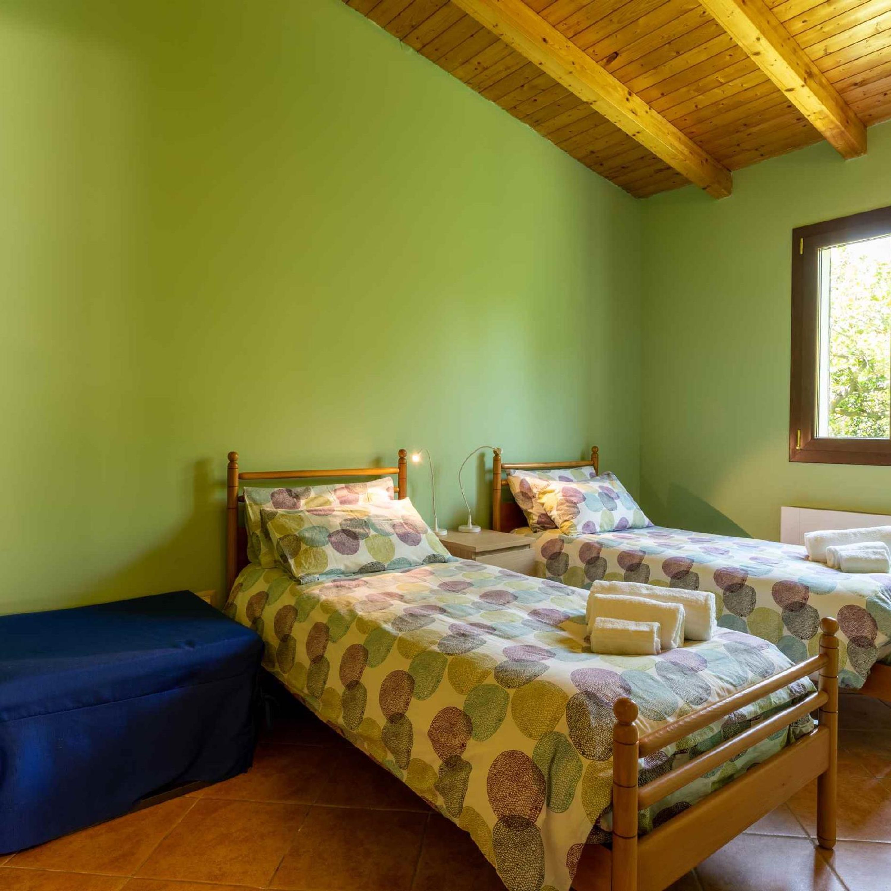 A room with two wooden beds, a blue bench, and a window with a view of greenery.