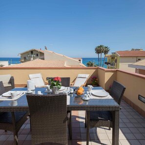 A dining area with wicker chairs and a table set for two, overlooking a coastal view with buildings and palm trees. - Apartment Eucaliptus sea View (Avola)