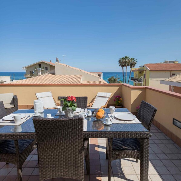 A dining area with wicker chairs and a table set for two, overlooking a coastal view with buildings and palm trees.