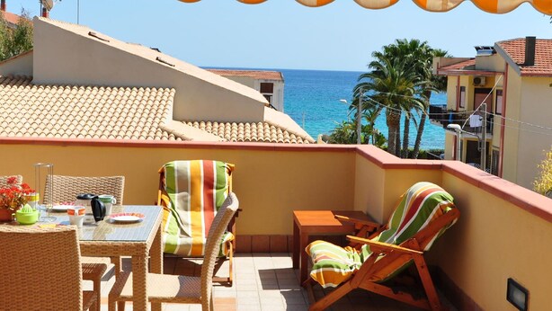 A balcony with a striped awning, wicker furniture, and a view of the sea. - Apartment Eucaliptus sea View (Avola)