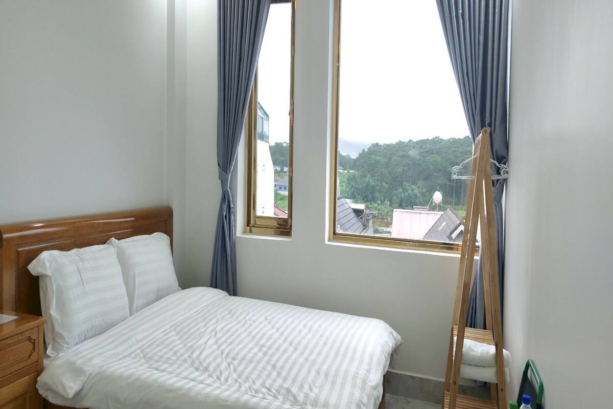 A bedroom with a wooden bed, two windows with curtains, a wooden ladder, and a view of buildings and greenery outside.