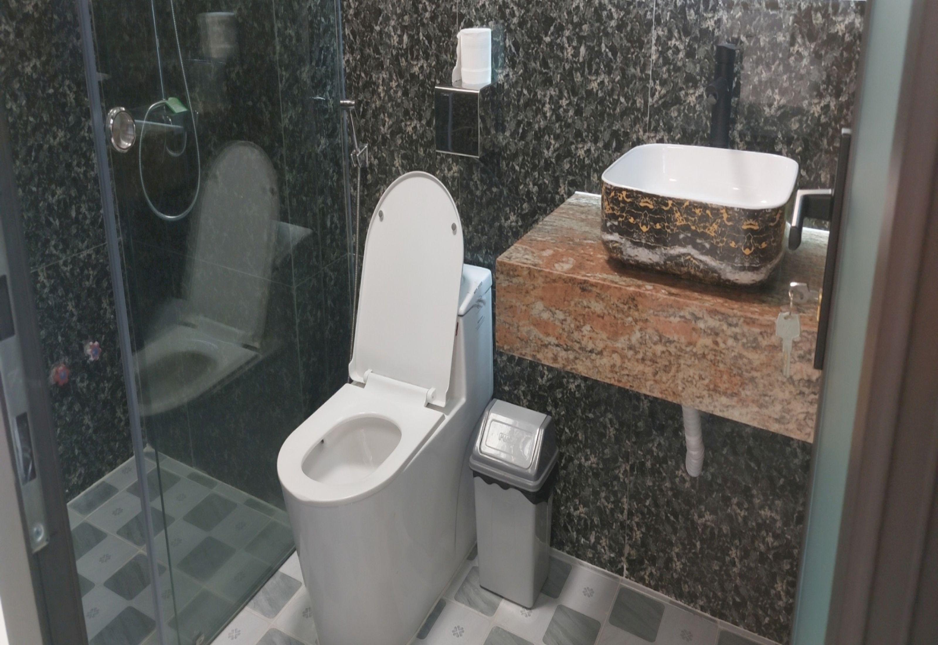 Um banheiro moderno com chuveiro em box de vidro, vaso sanitário, pia de mármore e lixeira.