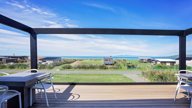 Una terraza de madera con un espacio de estar moderno al aire libre, vista despejada del océano y edificios distantes, y un césped bien cuidado.