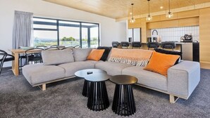 Una sala de estar moderna con un sofá gris, dos taburetes negros, techo de madera y una zona de cocina con sillas negras.