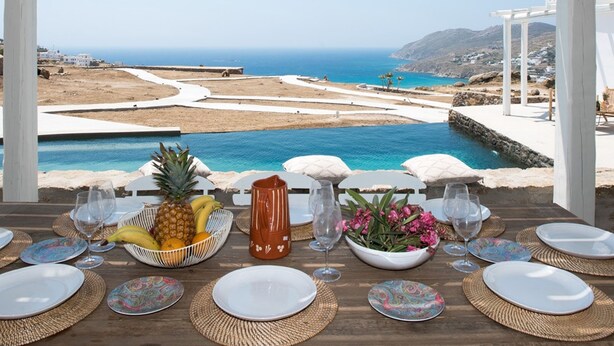 Mesa dispuesta para comer con vista al mar y un paisaje montañoso.