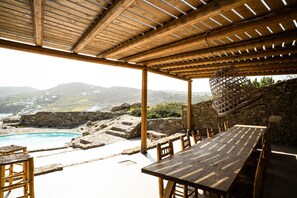 A wooden pergola with a table and chairs overlooking a swimming pool and mountainous landscape. - Villa Mykonos Kalo Livadi (Mykonos)