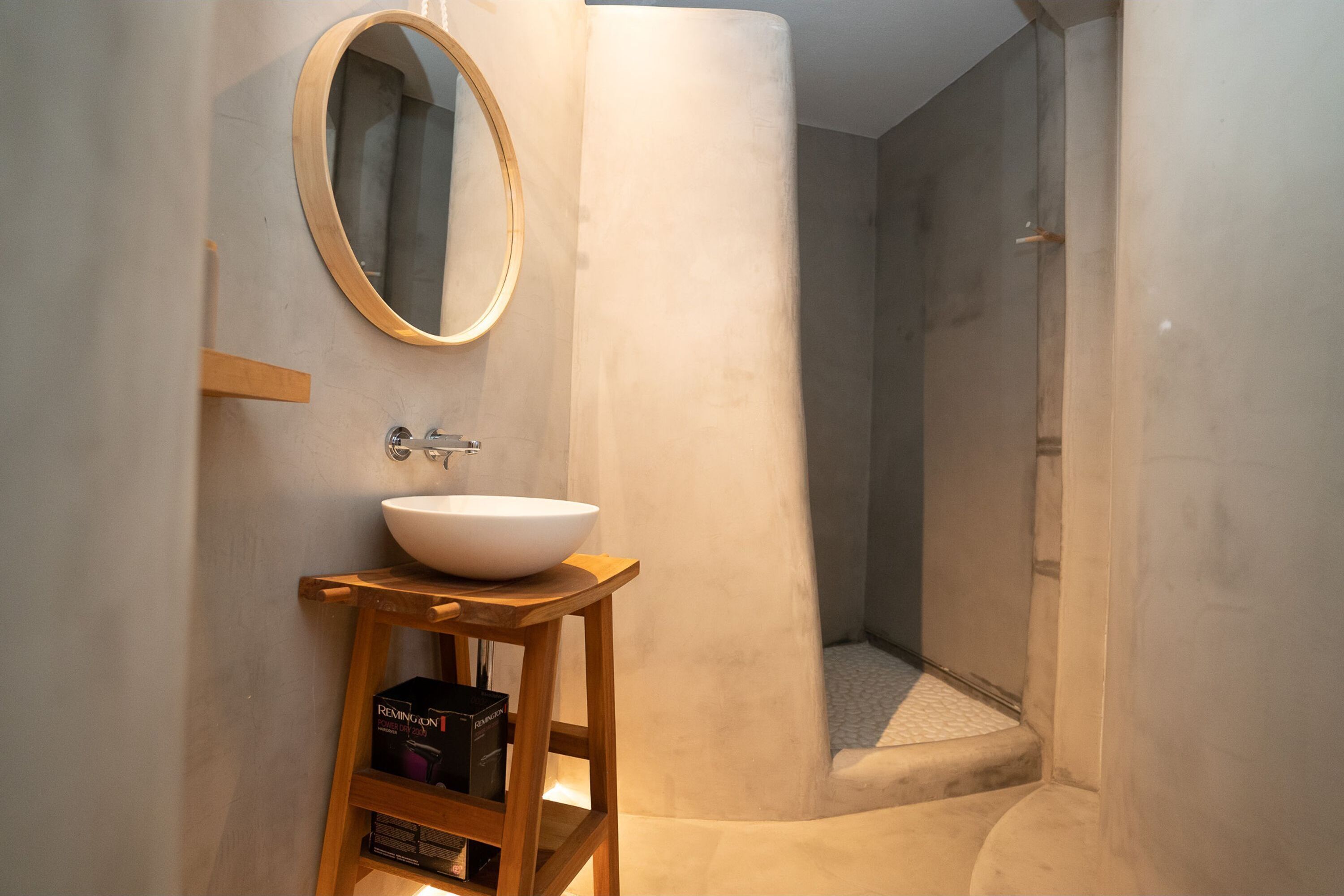 A bathroom with a round mirror, a white sink on a wooden stool, and a shower area.
