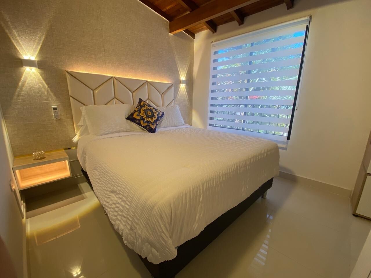 Une chambre moderne avec un grand lit, une tête de lit ornée de motifs géométriques, une table de chevet avec une lampe et une fenêtre avec des stores.