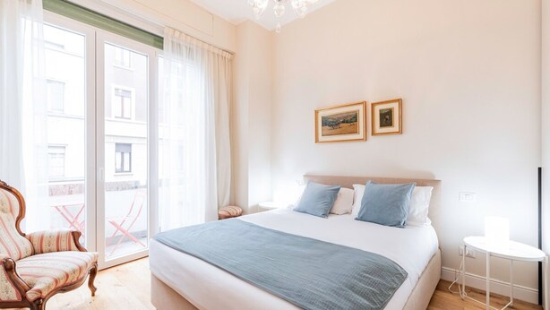 A hotel room with a large bed, a chair, a small table, and a balcony with a view of buildings. - Appartamento Tiepolo Elegant Venue in Porta Venezia (Milan)