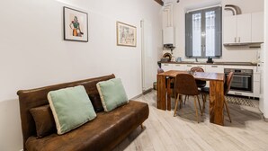 Un salon chaleureux avec un canapé en cuir marron, une table à manger en bois et des chaises. La cuisine est dotée de meubles blancs et d’une fenêtre située au-dessus de l’évier.