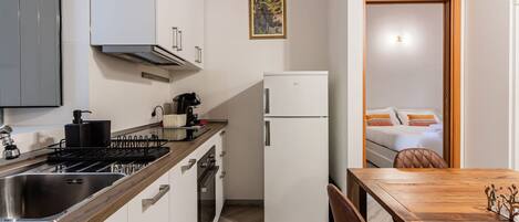 Cocina compacta con estufa, pileta y refrigerador blanco. Se ve una mesa de comedor con sillas, y una habitación con cama y ventana a través de una puerta abierta.