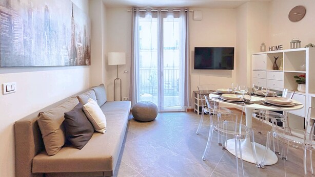 A modern living room with a sofa, dining table, and chairs, a flat-screen TV, and a large window with curtains. - Moderno Bilocale con Terrazzo e Parcheggio (Florence)