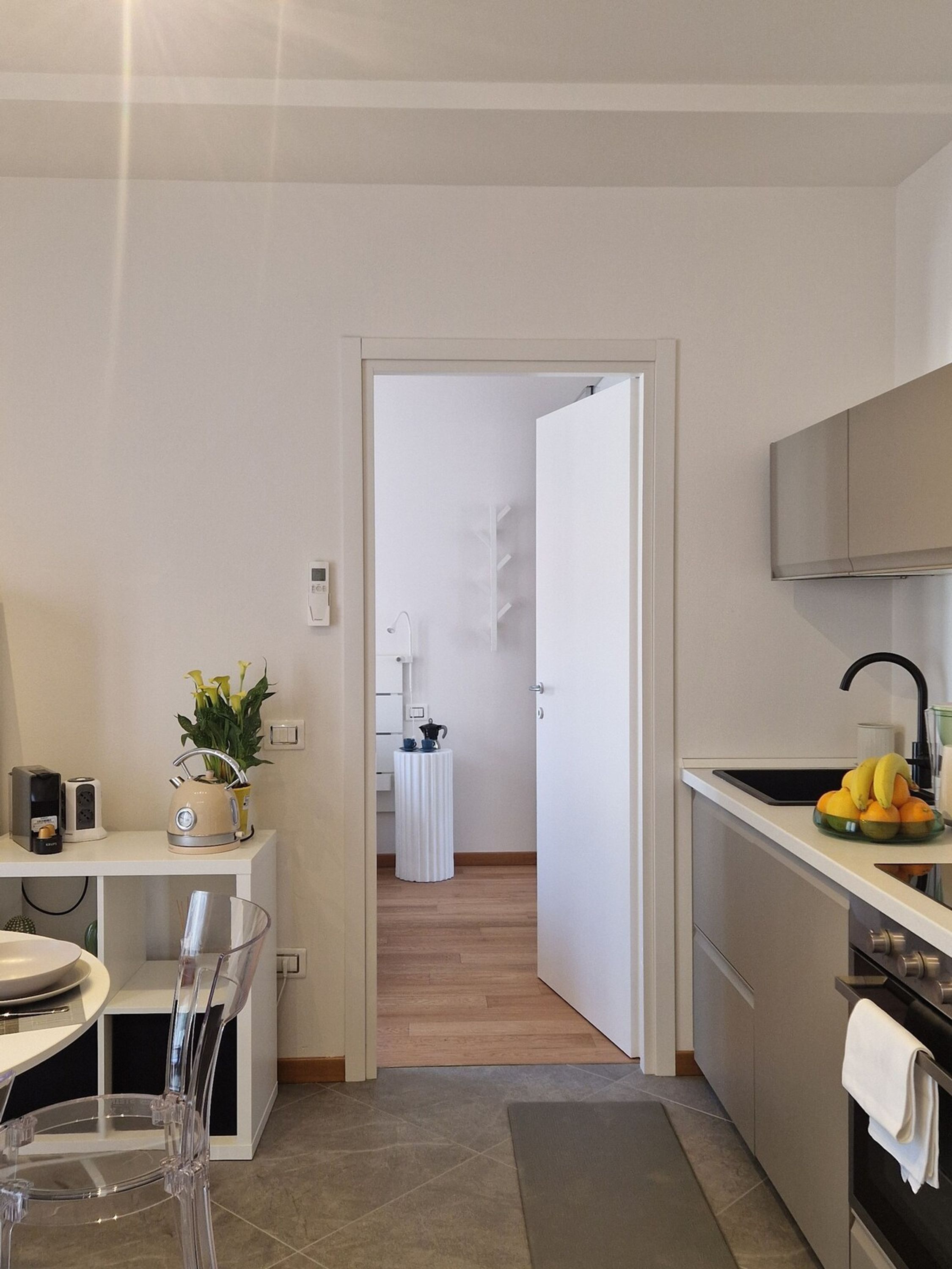 A modern kitchen with a white countertop, a sink, and a stove. There is a dining area with a clear glass table and chairs. A small white table with a plant and a kettle is visible in the background.