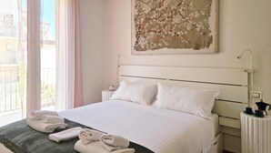 A neatly made bed with white linens, a green throw blanket, and rolled towels on the bed. There is a white nightstand with a lamp and a small table with a coffee pot and cups. A large painting of a tree is on the wall above the bed. - Moderno Bilocale con Terrazzo e Parcheggio (Florence)