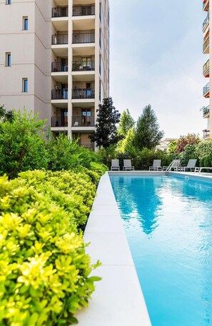 Piscina com espreguiçadeiras, rodeada por vegetação e adjacente a um edifício de vários andares.
