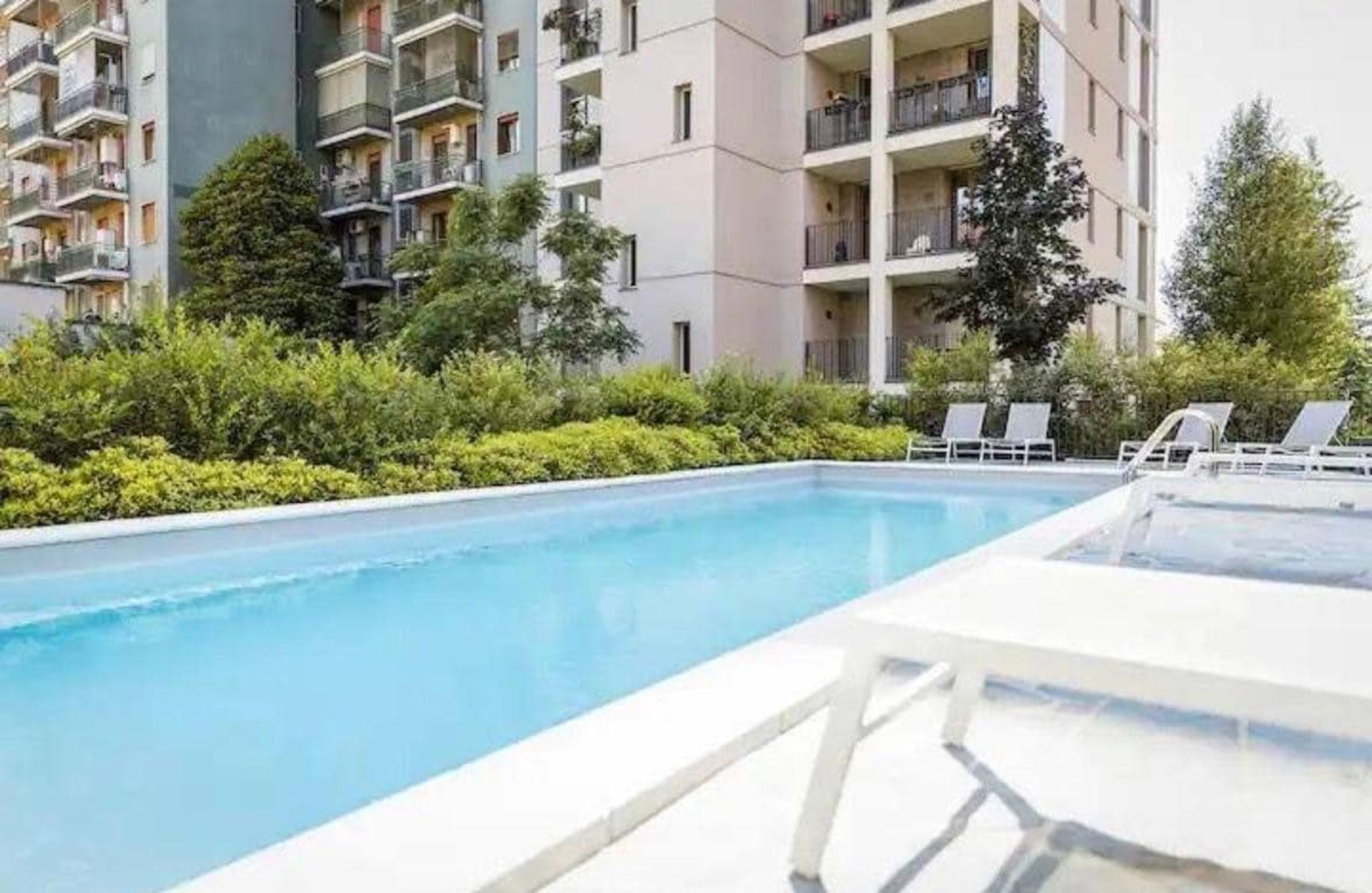 Une piscine entourée de chaises longues, bordée de verdure, et un immeuble à plusieurs étages doté de balcons.